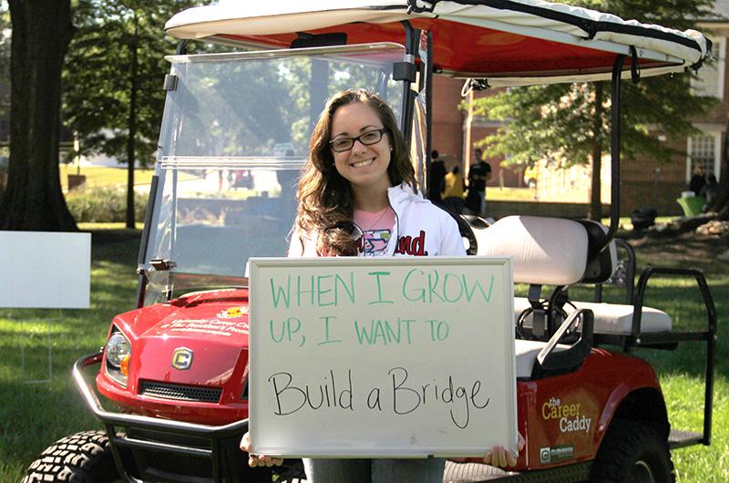 A person holding a whiteboard that reads "When I grow up, I want to Build a Bridge" in front of a red golf cart.